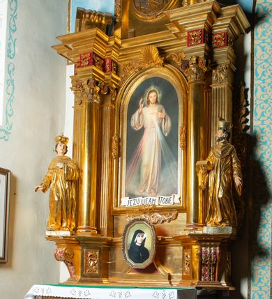 Zdjęcie nr 34: Nastawa architektoniczna, jednoosiowa, dwukondygnacyjna. Ołtarz murowany, prostopadłościenny, obity metalową blachą, zdobioną trybowaną kratownicą i gwiazdkami. Wewnątrz grób Chrystusa ze współczesną rzeźbą. Nastawa na niskim cokole, z wyodrębnionymi dwoma, ukośnie ustawionymi prostopadłościanami, z konsolami, dekorowanymi akantem i kampanulami. Pośrodku cokołu zamontowany obraz św. Faustyny w kształcie wieloboku. Kondygnacja flankowana dwiema parami pilastrów o kanelowanych trzonach oraz ustawioną przed nimi parą kolumn o kapitelach kompozytowych. Podpory dźwigają pełne belkowanie, wyłamane na ich osi, z silnie wysuniętym gzymsem, z przerwanym architrawem i fryzem nad polem środkowym. Na belkowaniu fragmenty przerwanego, wolutowego przyczółka, zwrócone pod kątem, do wewnątrz. W polu środkowym wnęka w formie stojącego prostokąta zamkniętego łukiem półkolistym, ujęta profilowaną ramą dekorowaną na bokach kwiatami, zwieńczoną muszlą. We wnęce obraz „Jezu Ufam Tobie”. Na konsolach, po bokach nastawy ustawione figury św. Kazimierza i św. Jana Kantego. Nastawa flankowana uszami utworzonymi z karbowanej wstęgi i ornamentu roślinnego. Druga kondygnacja węższa, flankowana ustawionymi pod kątem pilastrami, o trzonach dekorowanych kampanulami. Pilastry podtrzymują gzyms wyłamany na ich osi, nad polem przyjmujący kształt łuku półkolistego nadwieszonego. Na nim ustawione dwa wazony z kwiatami i bukiet kwiatów. W polu obraz św. Joachima w formie stojącego owalu, ujęty ramą, dekorowaną ornamentem roślinnym. Struktura niemal w całości złocona, elementy cokołu i fryzu polichromowane w kolorze czerwonym, ornamentyka złocona. 


