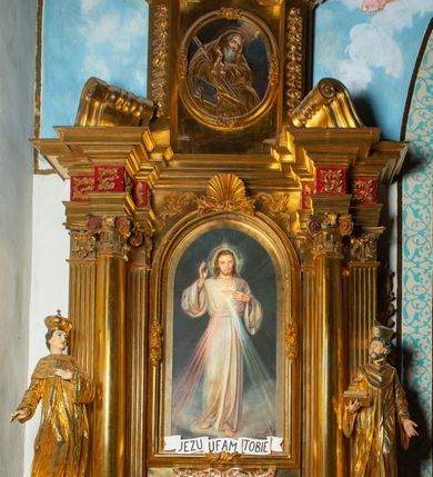 Zdjęcie nr 45: Nastawa architektoniczna, jednoosiowa, dwukondygnacyjna. Ołtarz murowany, prostopadłościenny, obity metalową blachą, zdobioną trybowaną kratownicą i gwiazdkami. Wewnątrz grób Chrystusa ze współczesną rzeźbą. Nastawa na niskim cokole, z wyodrębnionymi dwoma, ukośnie ustawionymi prostopadłościanami, z konsolami, dekorowanymi akantem i kampanulami. Pośrodku cokołu zamontowany obraz św. Faustyny w kształcie wieloboku. Kondygnacja flankowana dwiema parami pilastrów o kanelowanych trzonach oraz ustawioną przed nimi parą kolumn o kapitelach kompozytowych. Podpory dźwigają pełne belkowanie, wyłamane na ich osi, z silnie wysuniętym gzymsem, z przerwanym architrawem i fryzem nad polem środkowym. Na belkowaniu fragmenty przerwanego, wolutowego przyczółka, zwrócone pod kątem, do wewnątrz. W polu środkowym wnęka w formie stojącego prostokąta zamkniętego łukiem półkolistym, ujęta profilowaną ramą dekorowaną na bokach kwiatami, zwieńczoną muszlą. We wnęce obraz „Jezu Ufam Tobie”. Na konsolach, po bokach nastawy ustawione figury św. Kazimierza i św. Jana Kantego. Nastawa flankowana uszami utworzonymi z karbowanej wstęgi i ornamentu roślinnego. Druga kondygnacja węższa, flankowana ustawionymi pod kątem pilastrami, o trzonach dekorowanych kampanulami. Pilastry podtrzymują gzyms wyłamany na ich osi, nad polem przyjmujący kształt łuku półkolistego nadwieszonego. Na nim ustawione dwa wazony z kwiatami i bukiet kwiatów. W polu obraz św. Joachima w formie stojącego owalu, ujęty ramą, dekorowaną ornamentem roślinnym. Struktura niemal w całości złocona, elementy cokołu i fryzu polichromowane w kolorze czerwonym, ornamentyka złocona. 


