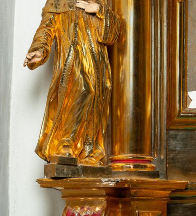 Zdjęcie nr 32: Nastawa architektoniczna, jednoosiowa, dwukondygnacyjna. Ołtarz murowany, prostopadłościenny, obity metalową blachą, zdobioną trybowaną kratownicą i gwiazdkami. Wewnątrz grób Chrystusa ze współczesną rzeźbą. Nastawa na niskim cokole, z wyodrębnionymi dwoma, ukośnie ustawionymi prostopadłościanami, z konsolami, dekorowanymi akantem i kampanulami. Pośrodku cokołu zamontowany obraz św. Faustyny w kształcie wieloboku. Kondygnacja flankowana dwiema parami pilastrów o kanelowanych trzonach oraz ustawioną przed nimi parą kolumn o kapitelach kompozytowych. Podpory dźwigają pełne belkowanie, wyłamane na ich osi, z silnie wysuniętym gzymsem, z przerwanym architrawem i fryzem nad polem środkowym. Na belkowaniu fragmenty przerwanego, wolutowego przyczółka, zwrócone pod kątem, do wewnątrz. W polu środkowym wnęka w formie stojącego prostokąta zamkniętego łukiem półkolistym, ujęta profilowaną ramą dekorowaną na bokach kwiatami, zwieńczoną muszlą. We wnęce obraz „Jezu Ufam Tobie”. Na konsolach, po bokach nastawy ustawione figury św. Kazimierza i św. Jana Kantego. Nastawa flankowana uszami utworzonymi z karbowanej wstęgi i ornamentu roślinnego. Druga kondygnacja węższa, flankowana ustawionymi pod kątem pilastrami, o trzonach dekorowanych kampanulami. Pilastry podtrzymują gzyms wyłamany na ich osi, nad polem przyjmujący kształt łuku półkolistego nadwieszonego. Na nim ustawione dwa wazony z kwiatami i bukiet kwiatów. W polu obraz św. Joachima w formie stojącego owalu, ujęty ramą, dekorowaną ornamentem roślinnym. Struktura niemal w całości złocona, elementy cokołu i fryzu polichromowane w kolorze czerwonym, ornamentyka złocona. 


