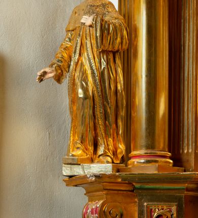 Zdjęcie nr 38: Nastawa architektoniczna, jednoosiowa, dwukondygnacyjna. Ołtarz murowany, prostopadłościenny, obity metalową blachą, zdobioną trybowaną kratownicą i gwiazdkami. Wewnątrz grób Chrystusa ze współczesną rzeźbą. Nastawa na niskim cokole, z wyodrębnionymi dwoma, ukośnie ustawionymi prostopadłościanami, z konsolami, dekorowanymi akantem i kampanulami. Pośrodku cokołu zamontowany obraz św. Faustyny w kształcie wieloboku. Kondygnacja flankowana dwiema parami pilastrów o kanelowanych trzonach oraz ustawioną przed nimi parą kolumn o kapitelach kompozytowych. Podpory dźwigają pełne belkowanie, wyłamane na ich osi, z silnie wysuniętym gzymsem, z przerwanym architrawem i fryzem nad polem środkowym. Na belkowaniu fragmenty przerwanego, wolutowego przyczółka, zwrócone pod kątem, do wewnątrz. W polu środkowym wnęka w formie stojącego prostokąta zamkniętego łukiem półkolistym, ujęta profilowaną ramą dekorowaną na bokach kwiatami, zwieńczoną muszlą. We wnęce obraz „Jezu Ufam Tobie”. Na konsolach, po bokach nastawy ustawione figury św. Kazimierza i św. Jana Kantego. Nastawa flankowana uszami utworzonymi z karbowanej wstęgi i ornamentu roślinnego. Druga kondygnacja węższa, flankowana ustawionymi pod kątem pilastrami, o trzonach dekorowanych kampanulami. Pilastry podtrzymują gzyms wyłamany na ich osi, nad polem przyjmujący kształt łuku półkolistego nadwieszonego. Na nim ustawione dwa wazony z kwiatami i bukiet kwiatów. W polu obraz św. Joachima w formie stojącego owalu, ujęty ramą, dekorowaną ornamentem roślinnym. Struktura niemal w całości złocona, elementy cokołu i fryzu polichromowane w kolorze czerwonym, ornamentyka złocona. 


