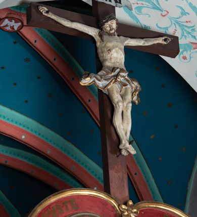 Zdjęcie nr 6: Drewniana belka o wklęsło-wypukłej formie utworzona z dwóch  stykających się wolut. Na jej froncie napis: „JEZU CHRYSTE ZMIŁUJ SIĘ NAD NAMI”. W centrum krzyż z figurą umarłego Chrystusa przybitą trzema gwoździami. Ciało w lekkim zwisie, o szeroko rozłożonych ramionach, z głową opadającą na prawy bark oraz stopami skrzyżowanymi w układzie prawa na lewą. Twarz ma szczupłą, o zamkniętych oczach, garbatym nosie i wąskich ustach, okoloną krótką brodą oraz kręconymi, brązowymi włosami opadającymi na ramiona. Na głowie ma koronę cierniową. Ciało jest wychudzone, o podkreślonych mięśniach ramion i nóg oraz żebrach klatki piersiowej. Przez biodra przewiązane krótkie, ekspresyjnie drapowane perizonium, rozwiane po bokach. Pionowa belka krzyża zwieńczona tabliczką z napisem „INRI”. 
Po lewej stronie krzyża figura Marii, ukazana frontalnie, w kontrapoście, pochylona, z dłońmi splecionymi z przodu. Twarz ma szczupłą, o małych oczach, prostym nosie i wąskich ustach. Ubrana jest w ciemnoczerwoną suknię, przepasaną w talii oraz niebieski płaszcz z jasnobrązową podszewką, zarzucony na lewe ramię i zawinięty wokół postaci. 
Po prawej stronie krzyża figura św. Jana, zwrócona w trzech czwartych w prawo, w kontrapoście, z uniesioną głową oraz rękami skierowanymi w prawo, z prawą w geście wskazującym. Twarz ma szczupłą, o oczach skierowanych w górę, prostym nosie i rozchylonych ustach, okoloną falowanymi włosami opadającymi na ramiona. Ubrany jest w jasnobrązową tunikę, przepasaną w talii oraz niebieski płaszcz z ciemnoczerwoną podszewką, zarzucony na prawe ramię i zawiązany na lewym boku. Polichromia ciał postaci naturalistyczna. 