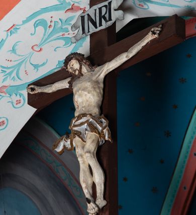 Zdjęcie nr 2: Drewniana belka o wklęsło-wypukłej formie utworzona z dwóch  stykających się wolut. Na jej froncie napis: „JEZU CHRYSTE ZMIŁUJ SIĘ NAD NAMI”. W centrum krzyż z figurą umarłego Chrystusa przybitą trzema gwoździami. Ciało w lekkim zwisie, o szeroko rozłożonych ramionach, z głową opadającą na prawy bark oraz stopami skrzyżowanymi w układzie prawa na lewą. Twarz ma szczupłą, o zamkniętych oczach, garbatym nosie i wąskich ustach, okoloną krótką brodą oraz kręconymi, brązowymi włosami opadającymi na ramiona. Na głowie ma koronę cierniową. Ciało jest wychudzone, o podkreślonych mięśniach ramion i nóg oraz żebrach klatki piersiowej. Przez biodra przewiązane krótkie, ekspresyjnie drapowane perizonium, rozwiane po bokach. Pionowa belka krzyża zwieńczona tabliczką z napisem „INRI”. 
Po lewej stronie krzyża figura Marii, ukazana frontalnie, w kontrapoście, pochylona, z dłońmi splecionymi z przodu. Twarz ma szczupłą, o małych oczach, prostym nosie i wąskich ustach. Ubrana jest w ciemnoczerwoną suknię, przepasaną w talii oraz niebieski płaszcz z jasnobrązową podszewką, zarzucony na lewe ramię i zawinięty wokół postaci. 
Po prawej stronie krzyża figura św. Jana, zwrócona w trzech czwartych w prawo, w kontrapoście, z uniesioną głową oraz rękami skierowanymi w prawo, z prawą w geście wskazującym. Twarz ma szczupłą, o oczach skierowanych w górę, prostym nosie i rozchylonych ustach, okoloną falowanymi włosami opadającymi na ramiona. Ubrany jest w jasnobrązową tunikę, przepasaną w talii oraz niebieski płaszcz z ciemnoczerwoną podszewką, zarzucony na prawe ramię i zawiązany na lewym boku. Polichromia ciał postaci naturalistyczna. 