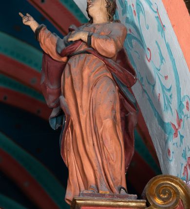 Zdjęcie nr 8: Drewniana belka o wklęsło-wypukłej formie utworzona z dwóch  stykających się wolut. Na jej froncie napis: „JEZU CHRYSTE ZMIŁUJ SIĘ NAD NAMI”. W centrum krzyż z figurą umarłego Chrystusa przybitą trzema gwoździami. Ciało w lekkim zwisie, o szeroko rozłożonych ramionach, z głową opadającą na prawy bark oraz stopami skrzyżowanymi w układzie prawa na lewą. Twarz ma szczupłą, o zamkniętych oczach, garbatym nosie i wąskich ustach, okoloną krótką brodą oraz kręconymi, brązowymi włosami opadającymi na ramiona. Na głowie ma koronę cierniową. Ciało jest wychudzone, o podkreślonych mięśniach ramion i nóg oraz żebrach klatki piersiowej. Przez biodra przewiązane krótkie, ekspresyjnie drapowane perizonium, rozwiane po bokach. Pionowa belka krzyża zwieńczona tabliczką z napisem „INRI”. 
Po lewej stronie krzyża figura Marii, ukazana frontalnie, w kontrapoście, pochylona, z dłońmi splecionymi z przodu. Twarz ma szczupłą, o małych oczach, prostym nosie i wąskich ustach. Ubrana jest w ciemnoczerwoną suknię, przepasaną w talii oraz niebieski płaszcz z jasnobrązową podszewką, zarzucony na lewe ramię i zawinięty wokół postaci. 
Po prawej stronie krzyża figura św. Jana, zwrócona w trzech czwartych w prawo, w kontrapoście, z uniesioną głową oraz rękami skierowanymi w prawo, z prawą w geście wskazującym. Twarz ma szczupłą, o oczach skierowanych w górę, prostym nosie i rozchylonych ustach, okoloną falowanymi włosami opadającymi na ramiona. Ubrany jest w jasnobrązową tunikę, przepasaną w talii oraz niebieski płaszcz z ciemnoczerwoną podszewką, zarzucony na prawe ramię i zawiązany na lewym boku. Polichromia ciał postaci naturalistyczna. 