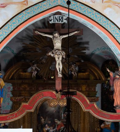 Zdjęcie nr 4: Drewniana belka o wklęsło-wypukłej formie utworzona z dwóch  stykających się wolut. Na jej froncie napis: „JEZU CHRYSTE ZMIŁUJ SIĘ NAD NAMI”. W centrum krzyż z figurą umarłego Chrystusa przybitą trzema gwoździami. Ciało w lekkim zwisie, o szeroko rozłożonych ramionach, z głową opadającą na prawy bark oraz stopami skrzyżowanymi w układzie prawa na lewą. Twarz ma szczupłą, o zamkniętych oczach, garbatym nosie i wąskich ustach, okoloną krótką brodą oraz kręconymi, brązowymi włosami opadającymi na ramiona. Na głowie ma koronę cierniową. Ciało jest wychudzone, o podkreślonych mięśniach ramion i nóg oraz żebrach klatki piersiowej. Przez biodra przewiązane krótkie, ekspresyjnie drapowane perizonium, rozwiane po bokach. Pionowa belka krzyża zwieńczona tabliczką z napisem „INRI”. 
Po lewej stronie krzyża figura Marii, ukazana frontalnie, w kontrapoście, pochylona, z dłońmi splecionymi z przodu. Twarz ma szczupłą, o małych oczach, prostym nosie i wąskich ustach. Ubrana jest w ciemnoczerwoną suknię, przepasaną w talii oraz niebieski płaszcz z jasnobrązową podszewką, zarzucony na lewe ramię i zawinięty wokół postaci. 
Po prawej stronie krzyża figura św. Jana, zwrócona w trzech czwartych w prawo, w kontrapoście, z uniesioną głową oraz rękami skierowanymi w prawo, z prawą w geście wskazującym. Twarz ma szczupłą, o oczach skierowanych w górę, prostym nosie i rozchylonych ustach, okoloną falowanymi włosami opadającymi na ramiona. Ubrany jest w jasnobrązową tunikę, przepasaną w talii oraz niebieski płaszcz z ciemnoczerwoną podszewką, zarzucony na prawe ramię i zawiązany na lewym boku. Polichromia ciał postaci naturalistyczna. 