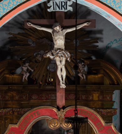 Zdjęcie nr 5: Drewniana belka o wklęsło-wypukłej formie utworzona z dwóch  stykających się wolut. Na jej froncie napis: „JEZU CHRYSTE ZMIŁUJ SIĘ NAD NAMI”. W centrum krzyż z figurą umarłego Chrystusa przybitą trzema gwoździami. Ciało w lekkim zwisie, o szeroko rozłożonych ramionach, z głową opadającą na prawy bark oraz stopami skrzyżowanymi w układzie prawa na lewą. Twarz ma szczupłą, o zamkniętych oczach, garbatym nosie i wąskich ustach, okoloną krótką brodą oraz kręconymi, brązowymi włosami opadającymi na ramiona. Na głowie ma koronę cierniową. Ciało jest wychudzone, o podkreślonych mięśniach ramion i nóg oraz żebrach klatki piersiowej. Przez biodra przewiązane krótkie, ekspresyjnie drapowane perizonium, rozwiane po bokach. Pionowa belka krzyża zwieńczona tabliczką z napisem „INRI”. 
Po lewej stronie krzyża figura Marii, ukazana frontalnie, w kontrapoście, pochylona, z dłońmi splecionymi z przodu. Twarz ma szczupłą, o małych oczach, prostym nosie i wąskich ustach. Ubrana jest w ciemnoczerwoną suknię, przepasaną w talii oraz niebieski płaszcz z jasnobrązową podszewką, zarzucony na lewe ramię i zawinięty wokół postaci. 
Po prawej stronie krzyża figura św. Jana, zwrócona w trzech czwartych w prawo, w kontrapoście, z uniesioną głową oraz rękami skierowanymi w prawo, z prawą w geście wskazującym. Twarz ma szczupłą, o oczach skierowanych w górę, prostym nosie i rozchylonych ustach, okoloną falowanymi włosami opadającymi na ramiona. Ubrany jest w jasnobrązową tunikę, przepasaną w talii oraz niebieski płaszcz z ciemnoczerwoną podszewką, zarzucony na prawe ramię i zawiązany na lewym boku. Polichromia ciał postaci naturalistyczna. 