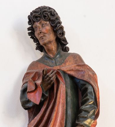 Zdjęcie nr 1: Pełnoplastyczna rzeźba ustawiona na niewielkiej, owalnej podstawie. Święty Jan przedstawiony całopostaciowo, w pozycji stojącej z ciałem zwróconym w trzech czwartych w prawo. Został ukazany jako szczupły mężczyzna o bujnych, kręconych włosach w kolorze ciemnobrązowym tworzących głęboko rzeźbione loki oraz chudej, „kościstej” twarzy z wyraźnie zaznaczonymi łukami brwiowymi i kośćmi policzkowymi. Wzrok uniesiony ku górze. Postać w lewej ręce trzyma księgę umieszczoną w brązowej sakiewce, prawą podtrzymuje połę płaszcza. Święty ubrany jest w ciemnozieloną, sięgającą ziemi szatę z długimi rękawami i wysokim kołnierzem oraz czerwony płaszcz. Spod szat widoczne są bose stopy. Figura w pełni polichromowana.