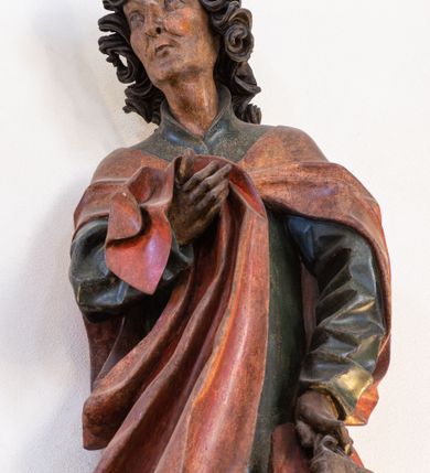 Zdjęcie nr 2: Pełnoplastyczna rzeźba ustawiona na niewielkiej, owalnej podstawie. Święty Jan przedstawiony całopostaciowo, w pozycji stojącej z ciałem zwróconym w trzech czwartych w prawo. Został ukazany jako szczupły mężczyzna o bujnych, kręconych włosach w kolorze ciemnobrązowym tworzących głęboko rzeźbione loki oraz chudej, „kościstej” twarzy z wyraźnie zaznaczonymi łukami brwiowymi i kośćmi policzkowymi. Wzrok uniesiony ku górze. Postać w lewej ręce trzyma księgę umieszczoną w brązowej sakiewce, prawą podtrzymuje połę płaszcza. Święty ubrany jest w ciemnozieloną, sięgającą ziemi szatę z długimi rękawami i wysokim kołnierzem oraz czerwony płaszcz. Spod szat widoczne są bose stopy. Figura w pełni polichromowana.