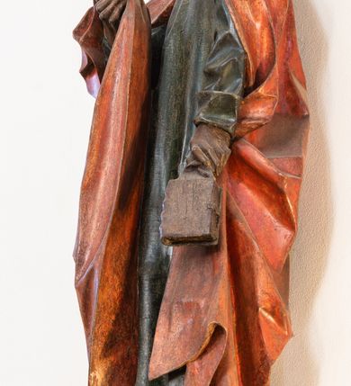 Zdjęcie nr 4: Pełnoplastyczna rzeźba ustawiona na niewielkiej, owalnej podstawie. Święty Jan przedstawiony całopostaciowo, w pozycji stojącej z ciałem zwróconym w trzech czwartych w prawo. Został ukazany jako szczupły mężczyzna o bujnych, kręconych włosach w kolorze ciemnobrązowym tworzących głęboko rzeźbione loki oraz chudej, „kościstej” twarzy z wyraźnie zaznaczonymi łukami brwiowymi i kośćmi policzkowymi. Wzrok uniesiony ku górze. Postać w lewej ręce trzyma księgę umieszczoną w brązowej sakiewce, prawą podtrzymuje połę płaszcza. Święty ubrany jest w ciemnozieloną, sięgającą ziemi szatę z długimi rękawami i wysokim kołnierzem oraz czerwony płaszcz. Spod szat widoczne są bose stopy. Figura w pełni polichromowana.