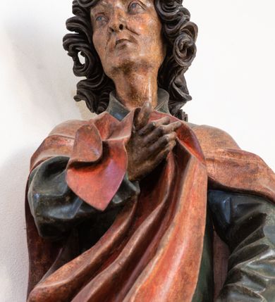 Zdjęcie nr 5: Pełnoplastyczna rzeźba ustawiona na niewielkiej, owalnej podstawie. Święty Jan przedstawiony całopostaciowo, w pozycji stojącej z ciałem zwróconym w trzech czwartych w prawo. Został ukazany jako szczupły mężczyzna o bujnych, kręconych włosach w kolorze ciemnobrązowym tworzących głęboko rzeźbione loki oraz chudej, „kościstej” twarzy z wyraźnie zaznaczonymi łukami brwiowymi i kośćmi policzkowymi. Wzrok uniesiony ku górze. Postać w lewej ręce trzyma księgę umieszczoną w brązowej sakiewce, prawą podtrzymuje połę płaszcza. Święty ubrany jest w ciemnozieloną, sięgającą ziemi szatę z długimi rękawami i wysokim kołnierzem oraz czerwony płaszcz. Spod szat widoczne są bose stopy. Figura w pełni polichromowana.