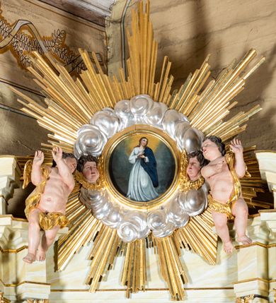 Zdjęcie nr 22: Ołtarz architektoniczny, jednokondygnacyjny, jednoosiowy ze zwieńczeniem i drewnianą, sarkofagową mensą. Na mensie ołtarza kremowe tabernakulum w formie prostopadłościennej szafki, ujęte parą jońskich, złoconych półkolumienek, stojących na profilowanym  cokoliku, wspierających belkowanie przechodzące nad polem centralnym w łuk. Drzwiczki w kształcie stojącego prostokąta zamkniętego łukiem pełnym zdobione płaskorzeźbionym motywem kielicha z hostią w glorii. Retabulum ołtarzowe na cokole, ujęte parą kolumn kompozytowych o spiralnie skręconych trzonach.  Podpory wspierają wyłamujące się ponad nimi oraz między nimi belkowanie o wysuniętym gzymsie, podtrzymujące przerwane fragmenty przyczółka. Pole środkowe w formie wnęki w kształcie stojącego prostokąta zamkniętego łukiem nadwieszonym z obrazem św. Anny Samotrzeć. Ujmuje ją para figur ustawionych na wysokich, zdwojonych cokołach. Postać z lewej strony trzyma berło, postać z prawej jabłko królewskie.  Zwieńczenie w formie stojącego owalu ujętego skłębionymi obłokami i szeroką glorią promienistą. W polu przedstawienie Matki Boskiej ujęte główkami anielskimi i parą putt. 
 Pola cokołu dekorowane płycinami. Na trzonach kolumn retabulum spiralnie oplatająca je wić roślinna. Wewnętrzne fragmenty wyłamanego gzymsu podtrzymują konsole, pod którymi podwieszono bukiety kwiatów. Struktura ołtarza polichromowana z imitacją marmoryzacji w odcieniach białym i kremowym, tło głowic kolumn czerwone, dekoracje i profile złocone i srebrzone. 

