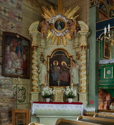 Zdjęcie nr 13: Ołtarz architektoniczny, jednokondygnacyjny, jednoosiowy ze zwieńczeniem i drewnianą, sarkofagową mensą. Na mensie ołtarza kremowe tabernakulum w formie prostopadłościennej szafki, ujęte parą jońskich, złoconych półkolumienek, stojących na profilowanym  cokoliku, wspierających belkowanie przechodzące nad polem centralnym w łuk. Drzwiczki w kształcie stojącego prostokąta zamkniętego łukiem pełnym zdobione płaskorzeźbionym motywem kielicha z hostią w glorii. Retabulum ołtarzowe na cokole, ujęte parą kolumn kompozytowych o spiralnie skręconych trzonach.  Podpory wspierają wyłamujące się ponad nimi oraz między nimi belkowanie o wysuniętym gzymsie, podtrzymujące przerwane fragmenty przyczółka. Pole środkowe w formie wnęki w kształcie stojącego prostokąta zamkniętego łukiem nadwieszonym z obrazem św. Anny Samotrzeć. Ujmuje ją para figur ustawionych na wysokich, zdwojonych cokołach. Postać z lewej strony trzyma berło, postać z prawej jabłko królewskie.  Zwieńczenie w formie stojącego owalu ujętego skłębionymi obłokami i szeroką glorią promienistą. W polu przedstawienie Matki Boskiej ujęte główkami anielskimi i parą putt. 
 Pola cokołu dekorowane płycinami. Na trzonach kolumn retabulum spiralnie oplatająca je wić roślinna. Wewnętrzne fragmenty wyłamanego gzymsu podtrzymują konsole, pod którymi podwieszono bukiety kwiatów. Struktura ołtarza polichromowana z imitacją marmoryzacji w odcieniach białym i kremowym, tło głowic kolumn czerwone, dekoracje i profile złocone i srebrzone. 

