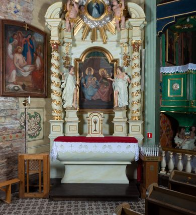 Zdjęcie nr 17: Ołtarz architektoniczny, jednokondygnacyjny, jednoosiowy ze zwieńczeniem i drewnianą, sarkofagową mensą. Na mensie ołtarza kremowe tabernakulum w formie prostopadłościennej szafki, ujęte parą jońskich, złoconych półkolumienek, stojących na profilowanym  cokoliku, wspierających belkowanie przechodzące nad polem centralnym w łuk. Drzwiczki w kształcie stojącego prostokąta zamkniętego łukiem pełnym zdobione płaskorzeźbionym motywem kielicha z hostią w glorii. Retabulum ołtarzowe na cokole, ujęte parą kolumn kompozytowych o spiralnie skręconych trzonach.  Podpory wspierają wyłamujące się ponad nimi oraz między nimi belkowanie o wysuniętym gzymsie, podtrzymujące przerwane fragmenty przyczółka. Pole środkowe w formie wnęki w kształcie stojącego prostokąta zamkniętego łukiem nadwieszonym z obrazem św. Anny Samotrzeć. Ujmuje ją para figur ustawionych na wysokich, zdwojonych cokołach. Postać z lewej strony trzyma berło, postać z prawej jabłko królewskie.  Zwieńczenie w formie stojącego owalu ujętego skłębionymi obłokami i szeroką glorią promienistą. W polu przedstawienie Matki Boskiej ujęte główkami anielskimi i parą putt. 
 Pola cokołu dekorowane płycinami. Na trzonach kolumn retabulum spiralnie oplatająca je wić roślinna. Wewnętrzne fragmenty wyłamanego gzymsu podtrzymują konsole, pod którymi podwieszono bukiety kwiatów. Struktura ołtarza polichromowana z imitacją marmoryzacji w odcieniach białym i kremowym, tło głowic kolumn czerwone, dekoracje i profile złocone i srebrzone. 
