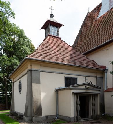 Zdjęcie nr 7: Kościół orientowany, murowany z cegły i kamienia, jednonawowy z trójprzęsłowym prezbiterium zamkniętym wielobocznie, sklepionym krzyżowo-żebrowo. Od północy umieszczona prostokątna, piętrowa zakrystia sklepiona kolebkowo (pięterko nad zakrystią dodane w 1885), dostępna kamiennym portalem o wykroju prostokątnym od strony wschodniej. Ściana północna zakrystii zakończona schodkowo z dekoracją blendową. Również od północy, na szerokości nawy do kościoła przylega prostokątna kaplica nakryta dachem czterospadowym zwieńczonym czworoboczną wieżyczką dostępna od zachodu przez prostokątną kruchtę z portalem o wykroju prostokątnym ujętym dwulalkowymi, kamiennymi balaskami. Kruchta zwieńczona trójkątnym przyczółkiem, na którym kamienny krzyż. Trójprzęsłowa nawa wyższa i szersza od prezbiterium i przesunięta w stosunku do jego osi ku stronie północnej, nakryta stropem. Północny mur nawy w części zachodniej poszerzony do pewnej wysokości dla pomieszczenia w jego grubości schodów prowadzących na chór muzyczny. Okna prostokątne zamknięte łukiem półkolistym. Zachodni i wschodni szczyt nawy zamknięty schodkowo. Od strony wschodniej trzy ostrołukowe blendy, w środkowej, wyżej od bocznych umieszczona tarcza herbowa z wizerunkiem św. Stanisława oraz Orła Białego. Od południa do nawy przylega prostokątna kruchta z kamiennym, ostrołukowym portalem nakryta dachem pulpitowym. Od strony zachodniej do bryły kościoła dostawiona prostokątna, współczesna kruchta wraz z chórem muzycznym (dobudowana w 1966) dostępna portalem o wykroju prostokątnym, nakryta dwuspadowym dachem. W ścianach bocznych wąskie, smukłe okna w kształcie stojącego prostokąta. Ponad kruchtą ażurowa dzwonnica na rzucie trójkąta zwieńczona krzyżem. Wieżyczka na sygnaturkę na rzucie ośmioboku zwieńczona barokowym hełmem z glorietą i krzyżem. Od zewnątrz kościół częściowo tynkowany, nawa i prezbiterium opięte szkarpami. Dachy kryte dachówką oraz blachą (część wschodnia prezbiterium).