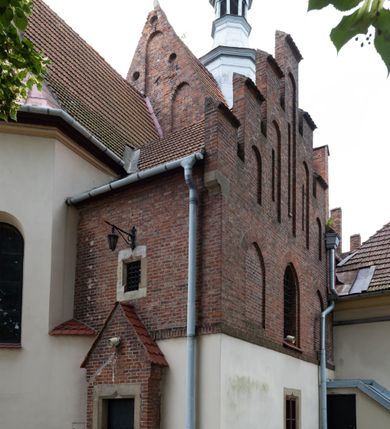 Zdjęcie nr 24: Kościół orientowany, murowany z cegły i kamienia, jednonawowy z trójprzęsłowym prezbiterium zamkniętym wielobocznie, sklepionym krzyżowo-żebrowo. Od północy umieszczona prostokątna, piętrowa zakrystia sklepiona kolebkowo (pięterko nad zakrystią dodane w 1885), dostępna kamiennym portalem o wykroju prostokątnym od strony wschodniej. Ściana północna zakrystii zakończona schodkowo z dekoracją blendową. Również od północy, na szerokości nawy do kościoła przylega prostokątna kaplica nakryta dachem czterospadowym zwieńczonym czworoboczną wieżyczką dostępna od zachodu przez prostokątną kruchtę z portalem o wykroju prostokątnym ujętym dwulalkowymi, kamiennymi balaskami. Kruchta zwieńczona trójkątnym przyczółkiem, na którym kamienny krzyż. Trójprzęsłowa nawa wyższa i szersza od prezbiterium i przesunięta w stosunku do jego osi ku stronie północnej, nakryta stropem. Północny mur nawy w części zachodniej poszerzony do pewnej wysokości dla pomieszczenia w jego grubości schodów prowadzących na chór muzyczny. Okna prostokątne zamknięte łukiem półkolistym. Zachodni i wschodni szczyt nawy zamknięty schodkowo. Od strony wschodniej trzy ostrołukowe blendy, w środkowej, wyżej od bocznych umieszczona tarcza herbowa z wizerunkiem św. Stanisława oraz Orła Białego. Od południa do nawy przylega prostokątna kruchta z kamiennym, ostrołukowym portalem nakryta dachem pulpitowym. Od strony zachodniej do bryły kościoła dostawiona prostokątna, współczesna kruchta wraz z chórem muzycznym (dobudowana w 1966) dostępna portalem o wykroju prostokątnym, nakryta dwuspadowym dachem. W ścianach bocznych wąskie, smukłe okna w kształcie stojącego prostokąta. Ponad kruchtą ażurowa dzwonnica na rzucie trójkąta zwieńczona krzyżem. Wieżyczka na sygnaturkę na rzucie ośmioboku zwieńczona barokowym hełmem z glorietą i krzyżem. Od zewnątrz kościół częściowo tynkowany, nawa i prezbiterium opięte szkarpami. Dachy kryte dachówką oraz blachą (część wschodnia prezbiterium).