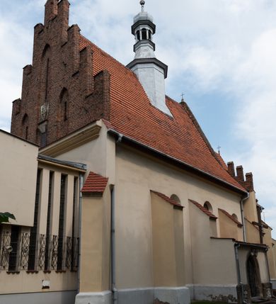 Zdjęcie nr 32: Kościół orientowany, murowany z cegły i kamienia, jednonawowy z trójprzęsłowym prezbiterium zamkniętym wielobocznie, sklepionym krzyżowo-żebrowo. Od północy umieszczona prostokątna, piętrowa zakrystia sklepiona kolebkowo (pięterko nad zakrystią dodane w 1885), dostępna kamiennym portalem o wykroju prostokątnym od strony wschodniej. Ściana północna zakrystii zakończona schodkowo z dekoracją blendową. Również od północy, na szerokości nawy do kościoła przylega prostokątna kaplica nakryta dachem czterospadowym zwieńczonym czworoboczną wieżyczką dostępna od zachodu przez prostokątną kruchtę z portalem o wykroju prostokątnym ujętym dwulalkowymi, kamiennymi balaskami. Kruchta zwieńczona trójkątnym przyczółkiem, na którym kamienny krzyż. Trójprzęsłowa nawa wyższa i szersza od prezbiterium i przesunięta w stosunku do jego osi ku stronie północnej, nakryta stropem. Północny mur nawy w części zachodniej poszerzony do pewnej wysokości dla pomieszczenia w jego grubości schodów prowadzących na chór muzyczny. Okna prostokątne zamknięte łukiem półkolistym. Zachodni i wschodni szczyt nawy zamknięty schodkowo. Od strony wschodniej trzy ostrołukowe blendy, w środkowej, wyżej od bocznych umieszczona tarcza herbowa z wizerunkiem św. Stanisława oraz Orła Białego. Od południa do nawy przylega prostokątna kruchta z kamiennym, ostrołukowym portalem nakryta dachem pulpitowym. Od strony zachodniej do bryły kościoła dostawiona prostokątna, współczesna kruchta wraz z chórem muzycznym (dobudowana w 1966) dostępna portalem o wykroju prostokątnym, nakryta dwuspadowym dachem. W ścianach bocznych wąskie, smukłe okna w kształcie stojącego prostokąta. Ponad kruchtą ażurowa dzwonnica na rzucie trójkąta zwieńczona krzyżem. Wieżyczka na sygnaturkę na rzucie ośmioboku zwieńczona barokowym hełmem z glorietą i krzyżem. Od zewnątrz kościół częściowo tynkowany, nawa i prezbiterium opięte szkarpami. Dachy kryte dachówką oraz blachą (część wschodnia prezbiterium).