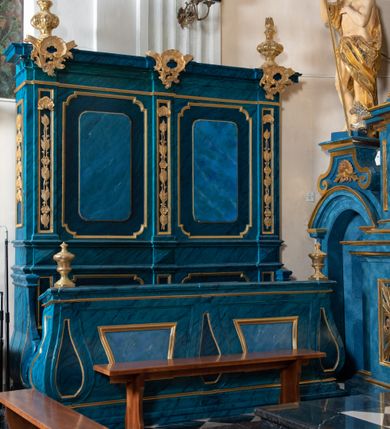 Zdjęcie nr 11: Dwie stalle o siedziskach obudowanych z przodu wybrzuszonymi ściankami z wazonami na pulpitach. Siedziska wybrzuszone z boku są ujęte falistymi poręczami, z plecami zdobionymi profilowanymi płycinami prostokątnymi o ściętych ćwierćkoliście narożach. Znajdujące się nad nimi wysokie zaplecki są ujęte pilastrami zdobionymi płycinami wypełnionymi wiciami liści i kwiatów z muszlami a z boku kampanuli, zwieńczone belkowaniem. W dwóch polach pomiędzy pilastrami zdwojone płyciny wyróżnione profilami w kształcie prostokątów o wyciętych ćwierćkoliście narożach.  Na belkowaniu trzy rokokowe kartusze i dwa płomieniste wazony. Polichromia imitująca jasno i ciemnoniebieską marmoryzację, ornamenty i profile złocone.  

Dwie stalle bez obudowy i wysokich zaplecków na falistych nogach, z których zewnętrzne poprzedzone lwimi łapami, z falistymi poręczami i prostymi oparciami zwieńczonymi gzymsem. Oparcie podzielone na trzy pola oddzielone żłobkowaniami i rautami nad nimi i wypełnione profilowanymi, kwadratowymi płycinami o ściętych ćwierćkoliście narożach.       