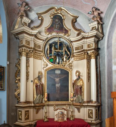 Zdjęcie nr 1: Przyścienna, jednokondygnacyjna, jednoosiowa nastawa ze zwieńczeniem uzyskała strukturę architektoniczną. Stipes ołtarzowy z drewnianą mensą z portatylem wykonano w kształcie sarkofagowym, ustawiając go na wysokim, fazowanym postumencie. Na nim znalazło się prostopadłościenne tabernakulum z obłym frontem udekorowanym prostokątnymi drzwiczkami zamkniętymi u góry nadwieszonym łukiem odcinkowy. Ich pole wypełnia płaskorzeźbione przedstawienie stojącego baranka apokaliptycznego. Całość wieńczy profilowany gzyms korrnujuacy utworzony z dwóch antetycznych wolut. Retabulum wsparto na dwuczęściowym, wysokim cokole. Kondygnację ujęto ustawionymi pod kątem opilastrowanymi, wklęsłymi filarami, przed którymi znalazła się para ustawionych pod kątem kolumn o kapitelach kompozytowych, usteweionyuch na postumentach o bokach dekorowanych prostokątnymi płycinami obramienionymi profilowanymi ramkami. Podpory dźwigają wyłamane imposty połączone ze sobą gzymsem. Na nim wsparto dwa falujące przyczółki, na których siedzi para putt. W polu kondygnacji umieszczono obraz Ukrzyżowania ujęty ramą w kształcie stojącego prostokąta zamkniętego u góry łukiem odcinkowym. Ponad nim na osi nastawę przepruto oculusem otwierającym się na okno witrażowe na wysokości przerwanego belkowania. Zwieńczenie ołtarza uzyskało kształt leżącego prostokąta o wklęsłych bokach utworzonych ze spływów wolutowych podtrzymujących profilowany gzyms koronujący powstały z dwóch antytetycznych wolut. W polu zwieńczenia umieszczono obraz św. Tytusa ujętego w fantazyjną ramę. Strukturę polichromowano na czarno. Głowice pilastrów i kapitele kolumn wyzłocono podobnie jak profile i obramowania. Rzeźby aniołków uzyskały naturalistyczną polichromię ze złoconymi przepaskami.   