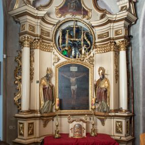 Zdjęcie nr 1: Przyścienna, jednokondygnacyjna, jednoosiowa nastawa ze zwieńczeniem uzyskała strukturę architektoniczną. Stipes ołtarzowy z drewnianą mensą z portatylem wykonano w kształcie sarkofagowym, ustawiając go na wysokim, fazowanym postumencie. Na nim znalazło się prostopadłościenne tabernakulum z obłym frontem udekorowanym prostokątnymi drzwiczkami zamkniętymi u góry nadwieszonym łukiem odcinkowy. Ich pole wypełnia płaskorzeźbione przedstawienie stojącego baranka apokaliptycznego. Całość wieńczy profilowany gzyms korrnujuacy utworzony z dwóch antetycznych wolut. Retabulum wsparto na dwuczęściowym, wysokim cokole. Kondygnację ujęto ustawionymi pod kątem opilastrowanymi, wklęsłymi filarami, przed którymi znalazła się para ustawionych pod kątem kolumn o kapitelach kompozytowych, usteweionyuch na postumentach o bokach dekorowanych prostokątnymi płycinami obramienionymi profilowanymi ramkami. Podpory dźwigają wyłamane imposty połączone ze sobą gzymsem. Na nim wsparto dwa falujące przyczółki, na których siedzi para putt. W polu kondygnacji umieszczono obraz Ukrzyżowania ujęty ramą w kształcie stojącego prostokąta zamkniętego u góry łukiem odcinkowym. Ponad nim na osi nastawę przepruto oculusem otwierającym się na okno witrażowe na wysokości przerwanego belkowania. Zwieńczenie ołtarza uzyskało kształt leżącego prostokąta o wklęsłych bokach utworzonych ze spływów wolutowych podtrzymujących profilowany gzyms koronujący powstały z dwóch antytetycznych wolut. W polu zwieńczenia umieszczono obraz św. Tytusa ujętego w fantazyjną ramę. Strukturę polichromowano na czarno. Głowice pilastrów i kapitele kolumn wyzłocono podobnie jak profile i obramowania. Rzeźby aniołków uzyskały naturalistyczną polichromię ze złoconymi przepaskami.   
