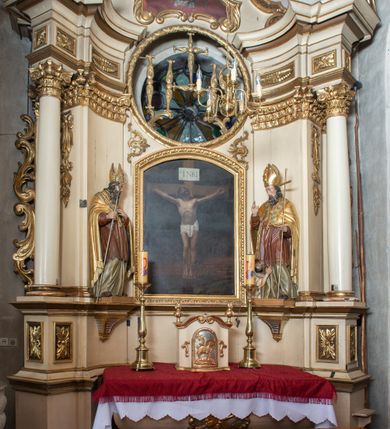 Zdjęcie nr 2: Przyścienna, jednokondygnacyjna, jednoosiowa nastawa ze zwieńczeniem uzyskała strukturę architektoniczną. Stipes ołtarzowy z drewnianą mensą z portatylem wykonano w kształcie sarkofagowym, ustawiając go na wysokim, fazowanym postumencie. Na nim znalazło się prostopadłościenne tabernakulum z obłym frontem udekorowanym prostokątnymi drzwiczkami zamkniętymi u góry nadwieszonym łukiem odcinkowy. Ich pole wypełnia płaskorzeźbione przedstawienie stojącego baranka apokaliptycznego. Całość wieńczy profilowany gzyms korrnujuacy utworzony z dwóch antetycznych wolut. Retabulum wsparto na dwuczęściowym, wysokim cokole. Kondygnację ujęto ustawionymi pod kątem opilastrowanymi, wklęsłymi filarami, przed którymi znalazła się para ustawionych pod kątem kolumn o kapitelach kompozytowych, usteweionyuch na postumentach o bokach dekorowanych prostokątnymi płycinami obramienionymi profilowanymi ramkami. Podpory dźwigają wyłamane imposty połączone ze sobą gzymsem. Na nim wsparto dwa falujące przyczółki, na których siedzi para putt. W polu kondygnacji umieszczono obraz Ukrzyżowania ujęty ramą w kształcie stojącego prostokąta zamkniętego u góry łukiem odcinkowym. Ponad nim na osi nastawę przepruto oculusem otwierającym się na okno witrażowe na wysokości przerwanego belkowania. Zwieńczenie ołtarza uzyskało kształt leżącego prostokąta o wklęsłych bokach utworzonych ze spływów wolutowych podtrzymujących profilowany gzyms koronujący powstały z dwóch antytetycznych wolut. W polu zwieńczenia umieszczono obraz św. Tytusa ujętego w fantazyjną ramę. Strukturę polichromowano na czarno. Głowice pilastrów i kapitele kolumn wyzłocono podobnie jak profile i obramowania. Rzeźby aniołków uzyskały naturalistyczną polichromię ze złoconymi przepaskami.   