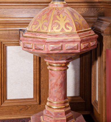 Zdjęcie nr 1: Chrzcielnica stojąca na ośmiobocznej dwustopniowej stopie. Ścianki stopy dekorowane rautami. Trzon w dolnej części o zdwojonym, ośmiobocznym cokole, wyżej w formie kolumny, zakończony półkolistym talerzykiem. Czara ośmioboczna, spłaszczona, każda ze ścianek dekorowana dwoma rautami. Pokrywa ośmioboczna w formie półkuli. Na szczycie ustawiona rzeźba św. Jana Chrzciciela. Święty ukazany w całej postaci, w delikatnym kontrapoście. W prawej dłoni trzyma dzbanuszek do polania wodą, a w lewej równoramienny krzyż na długim drzewcu. U jego prawej nogi leży biały baranek. Twarz podłużna o sumarycznie oddanych detalach, małe oczy i usta, krótki nos. Włosy bujne, ciemnobrązowe, opadają na plecy i ramiona, broda krótka rozdzielona na końcu w dwa pukle. Święty ubrany jest w suknię wykonaną z wielbłądziej skóry, sięgającą połowy łydek oraz czerwony płaszcz z jasnobeżową podszewką założony na prawe ramię i spływający pod lewą ręką. Draperia szat spokojna, niewielka. Polichromia w odsłoniętych partiach ciała naturalistyczna. Struktura chrzcielnicy marmoryzowana na kolor różowy, detale i profilowania złocone. 