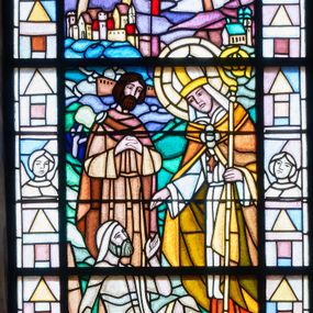 Zdjęcie nr 1: Witraż w kształcie stojącego prostokąta zamkniętego łukiem pełnym w szerokiej bordiurze wypełnionej popiersiami aniołów i figurami geometrycznymi. W centrum scena wskrzeszenia Piotrowina przez św. Stanisława. Po prawej stronie święty pochyla się nad zmartwychwstałym, wyciąga do niego prawą rękę, w lewej trzyma pastorał. Jest ubrany w czerwoną albę, białą komżę i obszerną żółtopomarańczową kapę spiętą na piersiach. Na głowie ma mitrę. Piotrowin siedzi w niszy grobowej wsparty na prawej ręce, lewą wyciąga w stronę świętego, lewą nogę ma zgiętą w kolanie. Biodra i głowę przykrywają mu jasne fragmenty całunu. Za nimi stoi męska postać z rękami złożonymi do modlitwy. Ma gęsty, ciemny zarost, jasnobrązową tunikę i ciemniejszy płaszcz spięty na prawym barku. Przed nimi fragment rozkopanej ziemi, tło stanowi krajobraz z łodzią na morzu oraz wybudowanymi na wzniesieniach miastem i kościołem z wieżami. W górnej partii tarcza herbowa otoczona od dołu wstęgą z napisem „1030 ŚW. STANISŁAW 1079”. W partii bordiury, przy dolnej krawędzi napisy „PROJEKTOWAŁ: / BOLESŁAW SZPECHT”, „WYKONALI: ANNA (dalej nieczytelne)”, „FUNDACJA / DR STANISŁAW / SĘK”. 