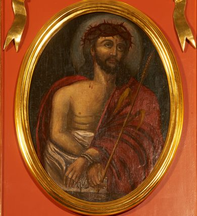 Zdjęcie nr 2: Obraz w kształcie stojącego owalu z przedstawieniem Chrystusa w typie Ecce Homo. Zbawiciel został ukazany w półpostaci, z rękami skrzyżowane przed sobą, związanymi i głową wzniesioną delikatnie w górę. Ma trójkątną twarz, szeroko otwarte oczy, uniesiony wzrok, wąski nos i usta. Twarz okala ciemny zarost, włosy opadają na barki i plecy, na głowie gruba korona cierniowa. Chrystus ma nagi tors i podkreślone żebra. Brzuch osłania biała tkanina, prawy bark i lewą rękę czerwony płaszcz. W lewej ręce trzyma trzcinę. Tło jednolite, ciemne, delikatnie rozjaśnione wokół głowy. 