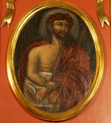 Zdjęcie nr 1: Obraz w kształcie stojącego owalu z przedstawieniem Chrystusa w typie Ecce Homo. Zbawiciel został ukazany w półpostaci, z rękami skrzyżowane przed sobą, związanymi i głową wzniesioną delikatnie w górę. Ma trójkątną twarz, szeroko otwarte oczy, uniesiony wzrok, wąski nos i usta. Twarz okala ciemny zarost, włosy opadają na barki i plecy, na głowie gruba korona cierniowa. Chrystus ma nagi tors i podkreślone żebra. Brzuch osłania biała tkanina, prawy bark i lewą rękę czerwony płaszcz. W lewej ręce trzyma trzcinę. Tło jednolite, ciemne, delikatnie rozjaśnione wokół głowy. 