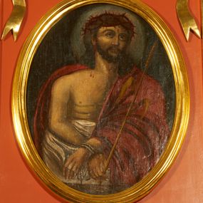 Zdjęcie nr 1: Obraz w kształcie stojącego owalu z przedstawieniem Chrystusa w typie Ecce Homo. Zbawiciel został ukazany w półpostaci, z rękami skrzyżowane przed sobą, związanymi i głową wzniesioną delikatnie w górę. Ma trójkątną twarz, szeroko otwarte oczy, uniesiony wzrok, wąski nos i usta. Twarz okala ciemny zarost, włosy opadają na barki i plecy, na głowie gruba korona cierniowa. Chrystus ma nagi tors i podkreślone żebra. Brzuch osłania biała tkanina, prawy bark i lewą rękę czerwony płaszcz. W lewej ręce trzyma trzcinę. Tło jednolite, ciemne, delikatnie rozjaśnione wokół głowy. 