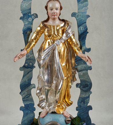 Zdjęcie nr 1: Postawa na planie prostokąta, obwiedziona profilowanym gzymsem, z otworami na drążki. Na niej ustawiona pełnoplastyczna figura Marii stojącej na globie oplecionym przez węża. Matka Boska ukazana frontalnie, z rękami rozłożonymi na boki, lewą stopą depcze głowę węża. Twarz ma owalną, o wysokim czole, wąskich oczach skierowanych w górę, krótkim nosie i wąskich ustach; okoloną długimi, brązowymi włosami, częściowo upiętymi z tyłu, a częściowo opadającymi na ramiona i kark. Ubrana jest w złoconą suknię, przepasaną w talii oraz srebrzony płaszcz przewiązany diagonalnie przez lewe ramię i opadający na plecy oraz prawą nogę. Polichromia ciała naturalistyczna. Nad figurą uproszczony czerwony baldachim z żółtym lambrekinem, wsparty na fantazyjnie wyciętych listewkach, polichromowanych razem z podstawą na sposób imitujący marmoryzację w kolorze niebiesko-szarym. 