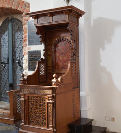 Zdjęcie nr 3: Konfesjonał typu architektonicznego na niskim cokole, o prostokątnym korpusie, ze zwieńczeniem w formie baldachimu. Na drzwiczkach płycina w kształcie zbliżonym do prostokąta, wypełniona symetrycznym ornamentem okuciowym z zaznaczonymi nitami, rozchodzącym się od centralnie umieszczonego rautu. Płycina jest flankowana tej samej wysokości pilastrami hermowymi z dekoracjami o motywach okuciowych i floralnych, które podtrzymują pełne belkowanie z fryzem z maureskową wicią roślinną. Ścianki boczne korpusu kontynuują podziały poziome drzwiczek. Są w przedniej części gładkie, a bliżej pleców dekorowane płycinami w kształcie stojącego prostokąta wypełnionymi czerwono-czarną maureską. Na lewej ściance dekoracja jest przesunięta ku przodowi i poprzedzona podziałami geometrycznymi, lecz jest częściowo ucięta. Powyżej poziomu drzwiczek kratownice, które sięgają połowie wysokości zaplecka i przechodzą ku frontowi w spływy wolutowe dochodzące do niewysokich cokołów z gzymsami i gałkami. Ścianki boczne zwężają się i rozszerzają w kierunku baldachimu ukazując szeroki, fantazyjnie wycięty, wklęsło-wypukły, zaczerniony profil. Przy tylnej krawędzi ścianki dekoruje wąski pas dzielonego na fragmenty ornamentu maureskowego, poniżej kratownicy na lewej ściance widoczny jest niewielki rysunek przypominjącym kiść owoców, kratownica na prawej ściance od dołu ujęta jest większym rysunkiem z motywem floralnym. Powyżej kratownic motyw okuciowy. Części dekoracji oddzielają szerokie, czarne ramy. Zaplecek  o rozbudowanej dekoracji - cokół dźwigający ujętą boniowaniem niszę flankowaną pilastrami podtrzymującymi pełne belkowanie. Płycina centralnej części cokołu wypełniona jest ornamentem rollwerkowym w kolorach czerwonym i zielonym oraz fragmentami czarnej wici maureskowej. Cokoły pilastrów plakietami z czerwoną maureską. W niszy rozbudowany ornament okuciowy utworzony z szerokich, czerwonych taśm z żółtymi nitami, przez który przewleczono czarną wić. Boniowanie ciemnobrązowe, dość silnie wysunięte. Pilastry o skomplikowanym kształcie, wklęsło-wypukłe w dolnej części, poprzedzielane fragmentami gzymsu, z równoległymi żłobieniami w kolorach żółtym i niebieskim, rautami na belkowaniu, wklęskami i fragmentami głowic inspirowanymi jońskimi. Na belkowaniu w części środkowej ornament okuciowy w kolorze czerwonym połączony z czarną wicia maureskową. Zwieńczenie w formie baldachimu złożonego z belkowania zakończonego profilowanym, silnie wysuniętym gzymsem. Fryz stanowią naprzemiennie ułożone większe fragmenty wici maureskowej i mniejsze, pojedyncze liście akantu. Zdobiony od dołu okuciami gzyms podtrzymują wklęsło-wypukłe kroksztyny, między którymi biegnie wąski pas ornamentu kostkowego. 