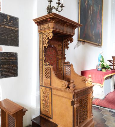 Zdjęcie nr 4: Konfesjonał typu architektonicznego na niskim cokole, o prostokątnym korpusie, ze zwieńczeniem w formie baldachimu. Na drzwiczkach płycina w kształcie zbliżonym do prostokąta, wypełniona symetrycznym ornamentem okuciowym z zaznaczonymi nitami, rozchodzącym się od centralnie umieszczonego rautu. Płycina jest flankowana tej samej wysokości pilastrami hermowymi z dekoracjami o motywach okuciowych i floralnych, które podtrzymują pełne belkowanie z fryzem z maureskową wicią roślinną. Ścianki boczne korpusu kontynuują podziały poziome drzwiczek. Są w przedniej części gładkie, a bliżej pleców dekorowane płycinami w kształcie stojącego prostokąta wypełnionymi czerwono-czarną maureską. Na lewej ściance dekoracja jest przesunięta ku przodowi i poprzedzona podziałami geometrycznymi, lecz jest częściowo ucięta. Powyżej poziomu drzwiczek kratownice, które sięgają połowie wysokości zaplecka i przechodzą ku frontowi w spływy wolutowe dochodzące do niewysokich cokołów z gzymsami i gałkami. Ścianki boczne zwężają się i rozszerzają w kierunku baldachimu ukazując szeroki, fantazyjnie wycięty, wklęsło-wypukły, zaczerniony profil. Przy tylnej krawędzi ścianki dekoruje wąski pas dzielonego na fragmenty ornamentu maureskowego, poniżej kratownicy na lewej ściance widoczny jest niewielki rysunek przypominjącym kiść owoców, kratownica na prawej ściance od dołu ujęta jest większym rysunkiem z motywem floralnym. Powyżej kratownic motyw okuciowy. Części dekoracji oddzielają szerokie, czarne ramy. Zaplecek  o rozbudowanej dekoracji - cokół dźwigający ujętą boniowaniem niszę flankowaną pilastrami podtrzymującymi pełne belkowanie. Płycina centralnej części cokołu wypełniona jest ornamentem rollwerkowym w kolorach czerwonym i zielonym oraz fragmentami czarnej wici maureskowej. Cokoły pilastrów plakietami z czerwoną maureską. W niszy rozbudowany ornament okuciowy utworzony z szerokich, czerwonych taśm z żółtymi nitami, przez który przewleczono czarną wić. Boniowanie ciemnobrązowe, dość silnie wysunięte. Pilastry o skomplikowanym kształcie, wklęsło-wypukłe w dolnej części, poprzedzielane fragmentami gzymsu, z równoległymi żłobieniami w kolorach żółtym i niebieskim, rautami na belkowaniu, wklęskami i fragmentami głowic inspirowanymi jońskimi. Na belkowaniu w części środkowej ornament okuciowy w kolorze czerwonym połączony z czarną wicia maureskową. Zwieńczenie w formie baldachimu złożonego z belkowania zakończonego profilowanym, silnie wysuniętym gzymsem. Fryz stanowią naprzemiennie ułożone większe fragmenty wici maureskowej i mniejsze, pojedyncze liście akantu. Zdobiony od dołu okuciami gzyms podtrzymują wklęsło-wypukłe kroksztyny, między którymi biegnie wąski pas ornamentu kostkowego. 
