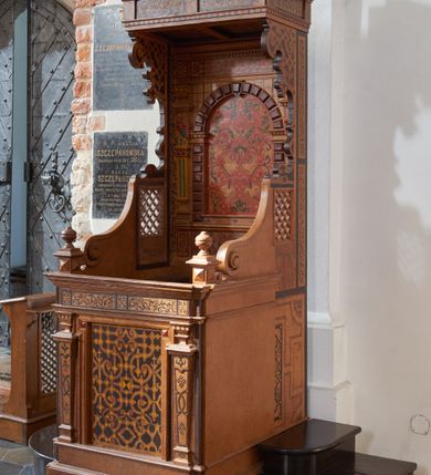 Zdjęcie nr 1: Konfesjonał typu architektonicznego na niskim cokole, o prostokątnym korpusie, ze zwieńczeniem w formie baldachimu. Na drzwiczkach płycina w kształcie zbliżonym do prostokąta, wypełniona symetrycznym ornamentem okuciowym z zaznaczonymi nitami, rozchodzącym się od centralnie umieszczonego rautu. Płycina jest flankowana tej samej wysokości pilastrami hermowymi z dekoracjami o motywach okuciowych i floralnych, które podtrzymują pełne belkowanie z fryzem z maureskową wicią roślinną. Ścianki boczne korpusu kontynuują podziały poziome drzwiczek. Są w przedniej części gładkie, a bliżej pleców dekorowane płycinami w kształcie stojącego prostokąta wypełnionymi czerwono-czarną maureską. Na lewej ściance dekoracja jest przesunięta ku przodowi i poprzedzona podziałami geometrycznymi, lecz jest częściowo ucięta. Powyżej poziomu drzwiczek kratownice, które sięgają połowie wysokości zaplecka i przechodzą ku frontowi w spływy wolutowe dochodzące do niewysokich cokołów z gzymsami i gałkami. Ścianki boczne zwężają się i rozszerzają w kierunku baldachimu ukazując szeroki, fantazyjnie wycięty, wklęsło-wypukły, zaczerniony profil. Przy tylnej krawędzi ścianki dekoruje wąski pas dzielonego na fragmenty ornamentu maureskowego, poniżej kratownicy na lewej ściance widoczny jest niewielki rysunek przypominjącym kiść owoców, kratownica na prawej ściance od dołu ujęta jest większym rysunkiem z motywem floralnym. Powyżej kratownic motyw okuciowy. Części dekoracji oddzielają szerokie, czarne ramy. Zaplecek  o rozbudowanej dekoracji - cokół dźwigający ujętą boniowaniem niszę flankowaną pilastrami podtrzymującymi pełne belkowanie. Płycina centralnej części cokołu wypełniona jest ornamentem rollwerkowym w kolorach czerwonym i zielonym oraz fragmentami czarnej wici maureskowej. Cokoły pilastrów plakietami z czerwoną maureską. W niszy rozbudowany ornament okuciowy utworzony z szerokich, czerwonych taśm z żółtymi nitami, przez który przewleczono czarną wić. Boniowanie ciemnobrązowe, dość silnie wysunięte. Pilastry o skomplikowanym kształcie, wklęsło-wypukłe w dolnej części, poprzedzielane fragmentami gzymsu, z równoległymi żłobieniami w kolorach żółtym i niebieskim, rautami na belkowaniu, wklęskami i fragmentami głowic inspirowanymi jońskimi. Na belkowaniu w części środkowej ornament okuciowy w kolorze czerwonym połączony z czarną wicia maureskową. Zwieńczenie w formie baldachimu złożonego z belkowania zakończonego profilowanym, silnie wysuniętym gzymsem. Fryz stanowią naprzemiennie ułożone większe fragmenty wici maureskowej i mniejsze, pojedyncze liście akantu. Zdobiony od dołu okuciami gzyms podtrzymują wklęsło-wypukłe kroksztyny, między którymi biegnie wąski pas ornamentu kostkowego. 