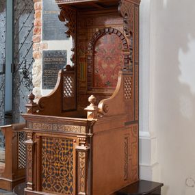 Zdjęcie nr 1: Konfesjonał typu architektonicznego na niskim cokole, o prostokątnym korpusie, ze zwieńczeniem w formie baldachimu. Na drzwiczkach płycina w kształcie zbliżonym do prostokąta, wypełniona symetrycznym ornamentem okuciowym z zaznaczonymi nitami, rozchodzącym się od centralnie umieszczonego rautu. Płycina jest flankowana tej samej wysokości pilastrami hermowymi z dekoracjami o motywach okuciowych i floralnych, które podtrzymują pełne belkowanie z fryzem z maureskową wicią roślinną. Ścianki boczne korpusu kontynuują podziały poziome drzwiczek. Są w przedniej części gładkie, a bliżej pleców dekorowane płycinami w kształcie stojącego prostokąta wypełnionymi czerwono-czarną maureską. Na lewej ściance dekoracja jest przesunięta ku przodowi i poprzedzona podziałami geometrycznymi, lecz jest częściowo ucięta. Powyżej poziomu drzwiczek kratownice, które sięgają połowie wysokości zaplecka i przechodzą ku frontowi w spływy wolutowe dochodzące do niewysokich cokołów z gzymsami i gałkami. Ścianki boczne zwężają się i rozszerzają w kierunku baldachimu ukazując szeroki, fantazyjnie wycięty, wklęsło-wypukły, zaczerniony profil. Przy tylnej krawędzi ścianki dekoruje wąski pas dzielonego na fragmenty ornamentu maureskowego, poniżej kratownicy na lewej ściance widoczny jest niewielki rysunek przypominjącym kiść owoców, kratownica na prawej ściance od dołu ujęta jest większym rysunkiem z motywem floralnym. Powyżej kratownic motyw okuciowy. Części dekoracji oddzielają szerokie, czarne ramy. Zaplecek  o rozbudowanej dekoracji - cokół dźwigający ujętą boniowaniem niszę flankowaną pilastrami podtrzymującymi pełne belkowanie. Płycina centralnej części cokołu wypełniona jest ornamentem rollwerkowym w kolorach czerwonym i zielonym oraz fragmentami czarnej wici maureskowej. Cokoły pilastrów plakietami z czerwoną maureską. W niszy rozbudowany ornament okuciowy utworzony z szerokich, czerwonych taśm z żółtymi nitami, przez który przewleczono czarną wić. Boniowanie ciemnobrązowe, dość silnie wysunięte. Pilastry o skomplikowanym kształcie, wklęsło-wypukłe w dolnej części, poprzedzielane fragmentami gzymsu, z równoległymi żłobieniami w kolorach żółtym i niebieskim, rautami na belkowaniu, wklęskami i fragmentami głowic inspirowanymi jońskimi. Na belkowaniu w części środkowej ornament okuciowy w kolorze czerwonym połączony z czarną wicia maureskową. Zwieńczenie w formie baldachimu złożonego z belkowania zakończonego profilowanym, silnie wysuniętym gzymsem. Fryz stanowią naprzemiennie ułożone większe fragmenty wici maureskowej i mniejsze, pojedyncze liście akantu. Zdobiony od dołu okuciami gzyms podtrzymują wklęsło-wypukłe kroksztyny, między którymi biegnie wąski pas ornamentu kostkowego. 