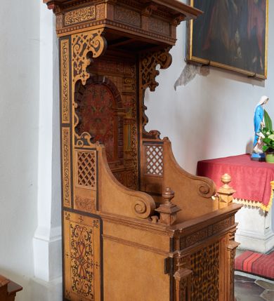 Zdjęcie nr 2: Konfesjonał typu architektonicznego na niskim cokole, o prostokątnym korpusie, ze zwieńczeniem w formie baldachimu. Na drzwiczkach płycina w kształcie zbliżonym do prostokąta, wypełniona symetrycznym ornamentem okuciowym z zaznaczonymi nitami, rozchodzącym się od centralnie umieszczonego rautu. Płycina jest flankowana tej samej wysokości pilastrami hermowymi z dekoracjami o motywach okuciowych i floralnych, które podtrzymują pełne belkowanie z fryzem z maureskową wicią roślinną. Ścianki boczne korpusu kontynuują podziały poziome drzwiczek. Są w przedniej części gładkie, a bliżej pleców dekorowane płycinami w kształcie stojącego prostokąta wypełnionymi czerwono-czarną maureską. Na lewej ściance dekoracja jest przesunięta ku przodowi i poprzedzona podziałami geometrycznymi, lecz jest częściowo ucięta. Powyżej poziomu drzwiczek kratownice, które sięgają połowie wysokości zaplecka i przechodzą ku frontowi w spływy wolutowe dochodzące do niewysokich cokołów z gzymsami i gałkami. Ścianki boczne zwężają się i rozszerzają w kierunku baldachimu ukazując szeroki, fantazyjnie wycięty, wklęsło-wypukły, zaczerniony profil. Przy tylnej krawędzi ścianki dekoruje wąski pas dzielonego na fragmenty ornamentu maureskowego, poniżej kratownicy na lewej ściance widoczny jest niewielki rysunek przypominjącym kiść owoców, kratownica na prawej ściance od dołu ujęta jest większym rysunkiem z motywem floralnym. Powyżej kratownic motyw okuciowy. Części dekoracji oddzielają szerokie, czarne ramy. Zaplecek  o rozbudowanej dekoracji - cokół dźwigający ujętą boniowaniem niszę flankowaną pilastrami podtrzymującymi pełne belkowanie. Płycina centralnej części cokołu wypełniona jest ornamentem rollwerkowym w kolorach czerwonym i zielonym oraz fragmentami czarnej wici maureskowej. Cokoły pilastrów plakietami z czerwoną maureską. W niszy rozbudowany ornament okuciowy utworzony z szerokich, czerwonych taśm z żółtymi nitami, przez który przewleczono czarną wić. Boniowanie ciemnobrązowe, dość silnie wysunięte. Pilastry o skomplikowanym kształcie, wklęsło-wypukłe w dolnej części, poprzedzielane fragmentami gzymsu, z równoległymi żłobieniami w kolorach żółtym i niebieskim, rautami na belkowaniu, wklęskami i fragmentami głowic inspirowanymi jońskimi. Na belkowaniu w części środkowej ornament okuciowy w kolorze czerwonym połączony z czarną wicia maureskową. Zwieńczenie w formie baldachimu złożonego z belkowania zakończonego profilowanym, silnie wysuniętym gzymsem. Fryz stanowią naprzemiennie ułożone większe fragmenty wici maureskowej i mniejsze, pojedyncze liście akantu. Zdobiony od dołu okuciami gzyms podtrzymują wklęsło-wypukłe kroksztyny, między którymi biegnie wąski pas ornamentu kostkowego. 