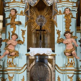 Zdjęcie nr 1: Tabernakulum pancerne, w formie drewnianej, prostopadłościennej skrzynki obitej blachą, o wypukłej ściance frontowej. Drzwiczki w formie kwadratu zamkniętego łukiem półkolistym, ujęte wzorzystym pasem, z trybowanym przedstawieniem baranka z krzyżem, leżącego na księdze, w glorii promienistej. W górnych narożach ścianki frontowej aplikowane elementy ornamentalne. Tabernakulum w drewnianej, architektonicznej obudowie, wpisane w cokół, na którym znajduje się tron wystawienia. Tron w formie niszy, ujęty dwiema parami zdwojonych pilastrów. Wewnętrza para wysunięta jest do przodu, a jej trzony dekorowane rocaille'm, kłosami zbóż, winogronami oraz główkami anielskimi przechodzą w spływy wolutowe, na których klęczą aniołki. Pilastry dźwigają belkowanie, wyłamane na ich osi, częściowo przerwane nad niszą środkową, z wyłamanym odcinkowo gzymsem, z podwieszoną tkaniną. Pilastry zewnętrzne ujęte wolutami spływającymi na wyłamane partie cokołu. Podstawa, pilastry i fryz dekorowane rocaille'm. Struktura polichromowana w kolorze błękitnym, ze złoconymi profilami i ornamentyką, tkanina srebrzona. 