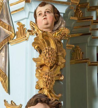Zdjęcie nr 6: Tabernakulum pancerne, w formie drewnianej, prostopadłościennej skrzynki obitej blachą, o wypukłej ściance frontowej. Drzwiczki w formie kwadratu zamkniętego łukiem półkolistym, ujęte wzorzystym pasem, z trybowanym przedstawieniem baranka z krzyżem, leżącego na księdze, w glorii promienistej. W górnych narożach ścianki frontowej aplikowane elementy ornamentalne. Tabernakulum w drewnianej, architektonicznej obudowie, wpisane w cokół, na którym znajduje się tron wystawienia. Tron w formie niszy, ujęty dwiema parami zdwojonych pilastrów. Wewnętrza para wysunięta jest do przodu, a jej trzony dekorowane rocaille'm, kłosami zbóż, winogronami oraz główkami anielskimi przechodzą w spływy wolutowe, na których klęczą aniołki. Pilastry dźwigają belkowanie, wyłamane na ich osi, częściowo przerwane nad niszą środkową, z wyłamanym odcinkowo gzymsem, z podwieszoną tkaniną. Pilastry zewnętrzne ujęte wolutami spływającymi na wyłamane partie cokołu. Podstawa, pilastry i fryz dekorowane rocaille'm. Struktura polichromowana w kolorze błękitnym, ze złoconymi profilami i ornamentyką, tkanina srebrzona. 