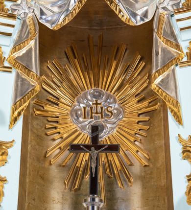 Zdjęcie nr 9: Tabernakulum pancerne, w formie drewnianej, prostopadłościennej skrzynki obitej blachą, o wypukłej ściance frontowej. Drzwiczki w formie kwadratu zamkniętego łukiem półkolistym, ujęte wzorzystym pasem, z trybowanym przedstawieniem baranka z krzyżem, leżącego na księdze, w glorii promienistej. W górnych narożach ścianki frontowej aplikowane elementy ornamentalne. Tabernakulum w drewnianej, architektonicznej obudowie, wpisane w cokół, na którym znajduje się tron wystawienia. Tron w formie niszy, ujęty dwiema parami zdwojonych pilastrów. Wewnętrza para wysunięta jest do przodu, a jej trzony dekorowane rocaille&#039;m, kłosami zbóż, winogronami oraz główkami anielskimi przechodzą w spływy wolutowe, na których klęczą aniołki. Pilastry dźwigają belkowanie, wyłamane na ich osi, częściowo przerwane nad niszą środkową, z wyłamanym odcinkowo gzymsem, z podwieszoną tkaniną. Pilastry zewnętrzne ujęte wolutami spływającymi na wyłamane partie cokołu. Podstawa, pilastry i fryz dekorowane rocaille&#039;m. Struktura polichromowana w kolorze błękitnym, ze złoconymi profilami i ornamentyką, tkanina srebrzona. 