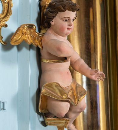 Zdjęcie nr 10: Tabernakulum pancerne, w formie drewnianej, prostopadłościennej skrzynki obitej blachą, o wypukłej ściance frontowej. Drzwiczki w formie kwadratu zamkniętego łukiem półkolistym, ujęte wzorzystym pasem, z trybowanym przedstawieniem baranka z krzyżem, leżącego na księdze, w glorii promienistej. W górnych narożach ścianki frontowej aplikowane elementy ornamentalne. Tabernakulum w drewnianej, architektonicznej obudowie, wpisane w cokół, na którym znajduje się tron wystawienia. Tron w formie niszy, ujęty dwiema parami zdwojonych pilastrów. Wewnętrza para wysunięta jest do przodu, a jej trzony dekorowane rocaille'm, kłosami zbóż, winogronami oraz główkami anielskimi przechodzą w spływy wolutowe, na których klęczą aniołki. Pilastry dźwigają belkowanie, wyłamane na ich osi, częściowo przerwane nad niszą środkową, z wyłamanym odcinkowo gzymsem, z podwieszoną tkaniną. Pilastry zewnętrzne ujęte wolutami spływającymi na wyłamane partie cokołu. Podstawa, pilastry i fryz dekorowane rocaille'm. Struktura polichromowana w kolorze błękitnym, ze złoconymi profilami i ornamentyką, tkanina srebrzona. 