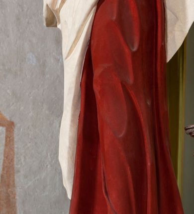 Zdjęcie nr 4: Rzeźba drążona z tyłu, ustawiona na niskiej podstawie. Figura ukazana frontalnie, z głową skierowaną w lewo, w kontrapoście, lewą ręką ociera chustą łzy, prawą kładzie na piersi. Święty Jan ma kwadratową twarz o odstających uszach, z wyodrębnionym podbródkiem, o małych, wąskich oczach, zmarszczonych brwiach, krótkim, garbatym nosie i małych, rozchylonych ustach z widoczną linią zębów. Włosy ma brązowe, krótkie i silnie kręcone, układające się na czubie i po bokach twarzy. Ubrany jest w czerwoną tunikę o niebieskiej podszewce, przepasaną w talii, ściśle przylegającą do ciała oraz biały płaszcz zawinięty wokół bioder. Polichromia ciała naturalistyczna. 