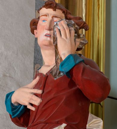 Zdjęcie nr 6: Rzeźba drążona z tyłu, ustawiona na niskiej podstawie. Figura ukazana frontalnie, z głową skierowaną w lewo, w kontrapoście, lewą ręką ociera chustą łzy, prawą kładzie na piersi. Święty Jan ma kwadratową twarz o odstających uszach, z wyodrębnionym podbródkiem, o małych, wąskich oczach, zmarszczonych brwiach, krótkim, garbatym nosie i małych, rozchylonych ustach z widoczną linią zębów. Włosy ma brązowe, krótkie i silnie kręcone, układające się na czubie i po bokach twarzy. Ubrany jest w czerwoną tunikę o niebieskiej podszewce, przepasaną w talii, ściśle przylegającą do ciała oraz biały płaszcz zawinięty wokół bioder. Polichromia ciała naturalistyczna. 