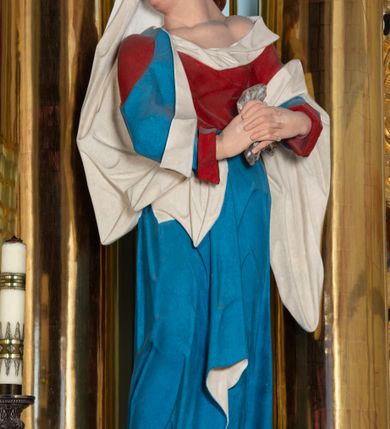 Zdjęcie nr 1: Rzeźba drążona z tyłu, ustawiona na niskiej podstawie. Figura ukazana frontalnie, z głową skierowaną w prawo, w kontrapoście, ze splecionymi z przodu dłońmi, w których trzyma chustę. Maria ma kwadratową twarz z odstającymi uszami, z wyodrębnionym podbródkiem, małymi, migdałowatymi oczami, krótkim nosem, niewielkimi, otwartymi ustami z widoczną linią zębów. Włosy ma brązowe, silnie kręcone, upięte z tyłu głowy. Ubrana jest w czerwoną, ściśle przylegającą tunikę, odsłaniającą lewe ramię oraz niebieski płaszcz o białej podszewce, zarzucony na ramiona i przewiązany przez biodra, ściśle opasujący ciało. Na głowie ma biały welon, zarzucony na lewą połowę głowy i podtrzymywany prawym ramieniem. Na stopach Maria ma brązowe sandały. Polichromia ciała jest naturalistyczna, o jasnej karnacji i zaróżowionych policzkach.