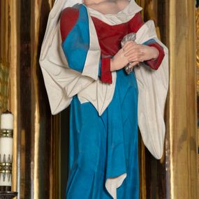 Zdjęcie nr 1: Rzeźba drążona z tyłu, ustawiona na niskiej podstawie. Figura ukazana frontalnie, z głową skierowaną w prawo, w kontrapoście, ze splecionymi z przodu dłońmi, w których trzyma chustę. Maria ma kwadratową twarz z odstającymi uszami, z wyodrębnionym podbródkiem, małymi, migdałowatymi oczami, krótkim nosem, niewielkimi, otwartymi ustami z widoczną linią zębów. Włosy ma brązowe, silnie kręcone, upięte z tyłu głowy. Ubrana jest w czerwoną, ściśle przylegającą tunikę, odsłaniającą lewe ramię oraz niebieski płaszcz o białej podszewce, zarzucony na ramiona i przewiązany przez biodra, ściśle opasujący ciało. Na głowie ma biały welon, zarzucony na lewą połowę głowy i podtrzymywany prawym ramieniem. Na stopach Maria ma brązowe sandały. Polichromia ciała jest naturalistyczna, o jasnej karnacji i zaróżowionych policzkach.