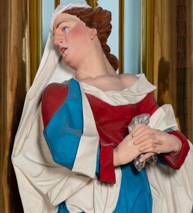 Zdjęcie nr 3: Rzeźba drążona z tyłu, ustawiona na niskiej podstawie. Figura ukazana frontalnie, z głową skierowaną w prawo, w kontrapoście, ze splecionymi z przodu dłońmi, w których trzyma chustę. Maria ma kwadratową twarz z odstającymi uszami, z wyodrębnionym podbródkiem, małymi, migdałowatymi oczami, krótkim nosem, niewielkimi, otwartymi ustami z widoczną linią zębów. Włosy ma brązowe, silnie kręcone, upięte z tyłu głowy. Ubrana jest w czerwoną, ściśle przylegającą tunikę, odsłaniającą lewe ramię oraz niebieski płaszcz o białej podszewce, zarzucony na ramiona i przewiązany przez biodra, ściśle opasujący ciało. Na głowie ma biały welon, zarzucony na lewą połowę głowy i podtrzymywany prawym ramieniem. Na stopach Maria ma brązowe sandały. Polichromia ciała jest naturalistyczna, o jasnej karnacji i zaróżowionych policzkach.