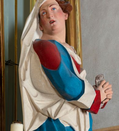Zdjęcie nr 5: Rzeźba drążona z tyłu, ustawiona na niskiej podstawie. Figura ukazana frontalnie, z głową skierowaną w prawo, w kontrapoście, ze splecionymi z przodu dłońmi, w których trzyma chustę. Maria ma kwadratową twarz z odstającymi uszami, z wyodrębnionym podbródkiem, małymi, migdałowatymi oczami, krótkim nosem, niewielkimi, otwartymi ustami z widoczną linią zębów. Włosy ma brązowe, silnie kręcone, upięte z tyłu głowy. Ubrana jest w czerwoną, ściśle przylegającą tunikę, odsłaniającą lewe ramię oraz niebieski płaszcz o białej podszewce, zarzucony na ramiona i przewiązany przez biodra, ściśle opasujący ciało. Na głowie ma biały welon, zarzucony na lewą połowę głowy i podtrzymywany prawym ramieniem. Na stopach Maria ma brązowe sandały. Polichromia ciała jest naturalistyczna, o jasnej karnacji i zaróżowionych policzkach.