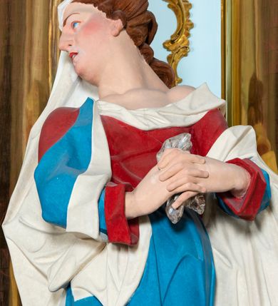Zdjęcie nr 7: Rzeźba drążona z tyłu, ustawiona na niskiej podstawie. Figura ukazana frontalnie, z głową skierowaną w prawo, w kontrapoście, ze splecionymi z przodu dłońmi, w których trzyma chustę. Maria ma kwadratową twarz z odstającymi uszami, z wyodrębnionym podbródkiem, małymi, migdałowatymi oczami, krótkim nosem, niewielkimi, otwartymi ustami z widoczną linią zębów. Włosy ma brązowe, silnie kręcone, upięte z tyłu głowy. Ubrana jest w czerwoną, ściśle przylegającą tunikę, odsłaniającą lewe ramię oraz niebieski płaszcz o białej podszewce, zarzucony na ramiona i przewiązany przez biodra, ściśle opasujący ciało. Na głowie ma biały welon, zarzucony na lewą połowę głowy i podtrzymywany prawym ramieniem. Na stopach Maria ma brązowe sandały. Polichromia ciała jest naturalistyczna, o jasnej karnacji i zaróżowionych policzkach.
