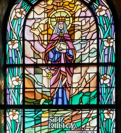 Zdjęcie nr 1: Witraż w kształcie stojącego prostokąta zamkniętego łukiem pełnym w szerokiej bordiurze wypełnionej wąskimi liśćmi i kwiatami lilii. W centrum kompozycji św. Kinga ukazana całopostaciowo, frontalnie. Lewą rękę ma wyprostowaną wzdłuż ciała, wypada z niej i leci w dół duży, złoty pierścień. Prawą rękę zgina na wysokości tułowia, w dłoni trzyma bryłę soli. Ma owalna twarz o surowych rysach, ciemne oczy, prosty nos i lekko rozchylone usta. Ubrana jest w długą, niebieską suknię oraz fioletowo-brązowy płaszcz, który ma zarzucony na ramiona, i którego końce przytrzymuje lewą dłonią. Głowę otaczają biała podwika i welon, na głowie otwarta, złota korona, wokół niej jasnożółty nimb. Pod postacią napis „ŚW. KINGA”, a pod nim mniejszymi literami: „FUNDATOR: STANISŁAW SĘK”. Na tej samej wysokości, z lewej strony „PROJEKTOWAŁ: / BOLESŁAW SZPRECHT / WYKONALI: / ANNA I IRENEUSZ ZAKRZYCCY / KRAKÓW 1999”. W partii bordiury otoczone ramką daty „1234-1292”. Tło abstrakcyjne, zielone w dolnej części, górą w odcieniach brązu. 