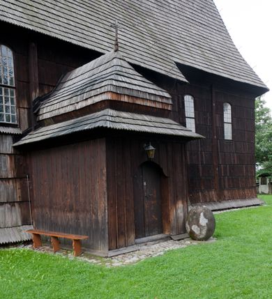 Zdjęcie nr 6: Kościół drewniany o konstrukcji zrębowej, elewacje jednolite, szalowane gontem. Wieża oraz kruchty zachodnia i południowa o konstrukcji słupowo-ramowej, kruchty szalowane deskami. Więźba dachowa drewniana, krokwiowa. Budynek na kamiennym podmurowaniu. 
Kościół orientowany, jednonawowy, nawa na rzucie prostokąta, prezbiterium węższe, wydłużone, zakończone trójbocznie. Do północnej ściany prezbiterium przylega zakrystia o wschodniej ścianie skośnej, będącej przedłużeniem bocznej ściany apsydy. 
Przy zachodniej ścianie nawy wieża na planie zbliżonym do kwadratu o wtórnym podziale na sale wystawiennicze (w przyziemiu wieży muzeum). Przy nawie od południa i przy wieży od frontu kruchty na planie kwadratu. 
Pomiędzy nawą a prezbiterium otwór tęczowy. 
Wnętrze nawy i prezbiterium nakryte pozornym sklepieniem kolebkowym, z zaskrzynieniami w nawie, podpartymi filarami o żłobkowanych trzonach, na cokołach, po trzy z każdej strony. Skrajem zaskrzynień nawy oraz górą ścian prezbiterium biegnie dekoracyjny gzyms drewniany, podparty w węgłach apsydy prezbiterialnej żłobkowanymi pilastrami. Łuk tęczowy półkolisty, wsparty na drewnianych konsolach. Otwory drzwiowe o prostokątnym wykroju zamknięte drzwiami obitymi deskami z metalowymi okuciami z prezbiterium do zakrystii, z nawy do kruchty południowej i z nawy do wieży. Okna w formie stojącego prostokąta zamkniętego łukiem odcinkowym, dwa w południowej ścianie prezbiterium, a w północnej dwa ślepe oraz cztery pary okien w północnej i południowej ścianie nawy. Na zamknięciu prezbiterium okulus z witrażem. Parapet chóru muzycznego w zachodniej części nawy, wsparty na dwóch drewnianych filarach, z wejściem z nawy po drabiniastych schodach z balustradą. Ściany i sklepienia pokryte polichromią z iluzjonistyczną architekturą i scenami figuralnymi. Posadzka kamienna.
Elewacja frontowa jednoosiowa. Na osi wejście do kruchty zachodniej. Korpus wieży o pochyłych ścianach, silnie zwężających się ku górze, zwieńczonych izbicą, ozdobioną u dołu koronką w formie zębów z frędzlami. Ściany kościoła, oprócz zakrystii i krucht szalowanych deskami, osłonięte rzędami gontów i opatrzone u dołu fartuchami. Na elewacji wschodniej ogrójec. 
Dach wyniosły, wspólny dla nawy i prezbiterium, hełm wieży cebulasty z latarnią, a w połowie długości dachu, nad nawą, wbudowana wieżyczka na sygnaturkę, wszystko kryte gontem. Kruchty nakryte osobnymi daszkami hełmowymi, baniastymi, krytymi gontem. 

Wokół kościoła rośnie starodrzew. Plac zamknięty kamiennym murem nakrytym drewnianym daszkiem dwuspadowym z 2 bramami i z 14 wieżyczkami (kapliczki-stacyjki) nakrytymi daszkami drewnianymi czterospadowymi. 