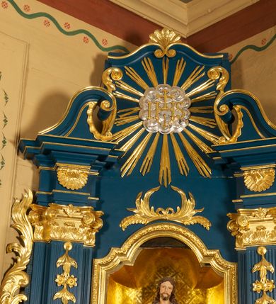Zdjęcie nr 2: Ołtarz prostopadłościenny, z drewnianą mensą. W polu antepedium trzy przedstawienia: Najświętszego Serca Marii, Świętej Rodziny oraz Najświętszego Serca Jezusa, każde ujęte złoconą ramą. 
Nastawa ołtarzowa jednoosiowa, jednokondygnacyjna ze zwieńczeniem, ustawiona na cokole. Kondygnacja nastawy ujęta zdwojonymi pilastrami o kapitelach kompozytowych i kanelowanych trzonach, ustawionych pod kątem do środka. Ustawione na wyodrębnionych cokołach podpory dźwigają imposty oraz fragmenty przerwanego przyczółka. W polu środkowym nisza w formie stojącego prostokąta zamkniętego łukiem półkolistym nadwieszonym z uskokiem, ujęta profilowaną ramą. Zwieńczenie w formie zbliżonej do rombu, ujętego spływami wolutowymi, zamkniętego stykającymi się wolutami. W polu rzeźbione przedstawienie monogramu IHS na obłokach, ujęte glorią promienistą. Cokoły pilastrów dekorowane płycinami utworzonymi z ornamentu roślinnego. W partii trzonów zwisy kwiatowe, a impostów palmety. Rama pola środkowego ujęta od gry i dołu ornamentem roślinnym. Nastawa flankowana uszami utworzonymi z rocaille'u i ornamentu roślinnego. Struktura w kolorze granatowym; ornamentyka oraz profile złocone, obłoki srebrzone.
