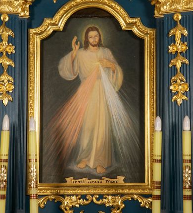 Zdjęcie nr 1: Obraz w kształcie stojącego prostokąta z wizerunkiem „Jezu ufam Tobie”. W centrum kompozycji całopostaciowe, przedstawienie Jezusa, ukazanego frontalnie, w wykroku,  prawą rękę unosi w geście błogosławieństwa, lewą składa na piersi, wskazując na miejsce serca, otoczone glorią promienistą, z którego wydobywają się wiązki promieni – czerwone na lewo, a białe na prawo. Na dłoniach i stopach widoczne ślady męki. Jezus ma owalną twarz z wysokim czołem, ciemnie oczy, wąski nos i małe usta. Jasnobrązowe włosy opadają mu na ramiona, ma krótkie wąsy i brodę dzielącą się w dwa pukle. Wokół głowy świetlisty nimb. Jest ubrany w białą, długą tunikę o szerokich rękawach, która jest przewiązana w pasie. Stoi na obłoku, na kamiennej posadzce. Wzdłuż dolnej krawędzi obrazu banderola z napisem: „JEZU UFAM TOBIE”. Tło obrazu jest jednolite, ciemne, a całość utrzymana w chłodnej kolorystyce. 
