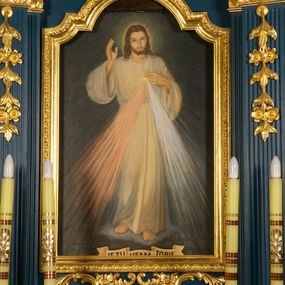 Zdjęcie nr 1: Obraz w kształcie stojącego prostokąta z wizerunkiem „Jezu ufam Tobie”. W centrum kompozycji całopostaciowe, przedstawienie Jezusa, ukazanego frontalnie, w wykroku,  prawą rękę unosi w geście błogosławieństwa, lewą składa na piersi, wskazując na miejsce serca, otoczone glorią promienistą, z którego wydobywają się wiązki promieni – czerwone na lewo, a białe na prawo. Na dłoniach i stopach widoczne ślady męki. Jezus ma owalną twarz z wysokim czołem, ciemnie oczy, wąski nos i małe usta. Jasnobrązowe włosy opadają mu na ramiona, ma krótkie wąsy i brodę dzielącą się w dwa pukle. Wokół głowy świetlisty nimb. Jest ubrany w białą, długą tunikę o szerokich rękawach, która jest przewiązana w pasie. Stoi na obłoku, na kamiennej posadzce. Wzdłuż dolnej krawędzi obrazu banderola z napisem: „JEZU UFAM TOBIE”. Tło obrazu jest jednolite, ciemne, a całość utrzymana w chłodnej kolorystyce. 